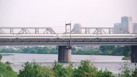 Shinkansen-Hochgeschwindigkeitszug-Fährt-Tagsüber-über-Die-Brücke-Eines-Ruhigen-Tamagawa-Flusses-Mit-Stadtlandschaft-Im-Hintergrund-In-Tokio,-Japan