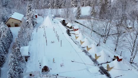 Aerial-view-towards-cottages,-sauna-and-a-big-tent-in-a-Finnish-holiday-resort-village,-in-middle-of-polar-forest,-on-a-cloudy,-winter-day---circling,-drone-shot