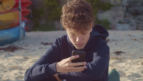 Teenage-boy-texts-on-smartphone-on-beach,-portrait