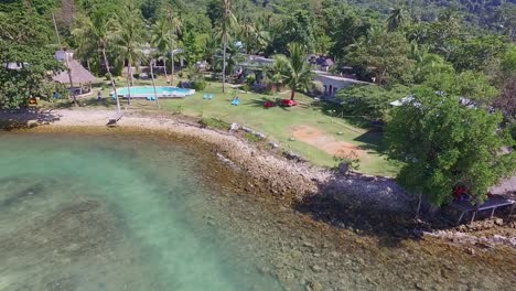 Panning-out-Drone-shot-of-small-bungalow-resort-in-Thailand-with-swimming-pool-and-surrounding-ocean-and-coral-reef