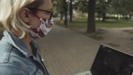 Side-View-Of-Caucasian-Lady-With-Eyeglasses-And-Mask-Using-Laptop-While-Sitting-At-The-Park---close-up