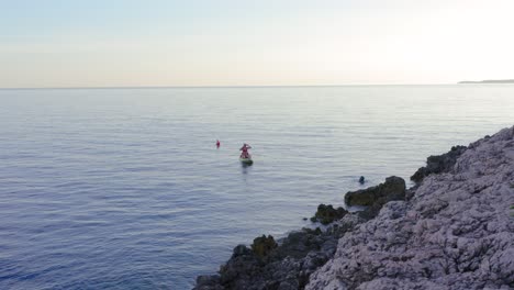 Mädchen-Sitzt-Und-Rudert-Kajak-Mit-Einem-Mann,-Der-In-Der-Nähe-Der-Felsigen-Küste-Der-Insel-Losinj-In-Kroatien-Schwimmt