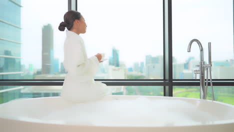 Asian-woman-sitting-on-resort-style-bathtub,-drinking-tea-and-looks-out-on-a-modern-skyline