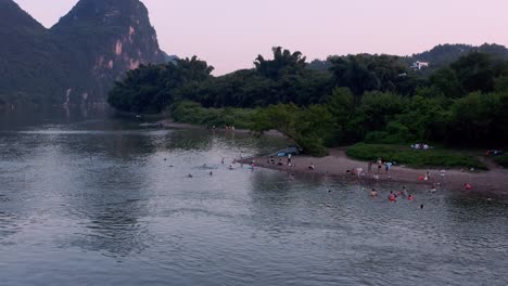 People-bathing-on-secluded-river-beach,-karst-mountain-China-landscape,-aerial