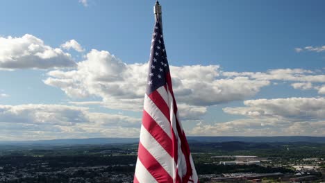 American-flag-close-up-aerial,-USA-pride,-Old-Glory-waves-in-wind-on-sunny-summer-day,-urban-city-town-center-in-valley-below-in-distance,-pulll-back-reveals-wide-panorama