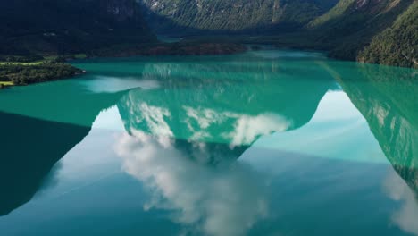 Moving-drone-shot-of-a-green-transparent-natural-lake-reflecting-the-mountains-and-clouds