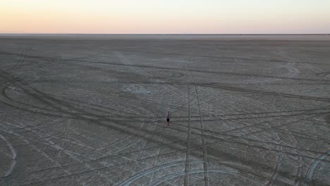 Lonely-Man-Standing-in-Twilight,-Endless-Landscape-of-Salt-Flats-With-Car-Trails