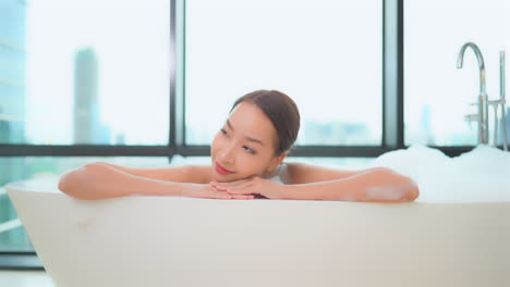 Asian-woman-relaxing-on-resort-style-bathtub,-modern-skyline-on-background