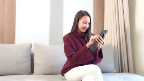 Junge-Hübsche-Asiatische-Frau,-Die-Auf-Dem-Sofa-Im-Wohnzimmer-SMS-Schreibt-Und-Auf-Dem-Smartphone-Chattet