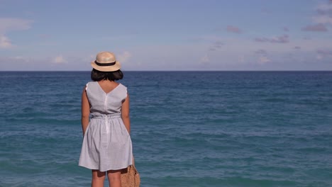 Back-of-girl-in-summer-dress-standing-in-front-of-wide-open-Ocean-Horizon-on-bright-and-sunny-day-with-high-waves-splashing-in-front-of-her