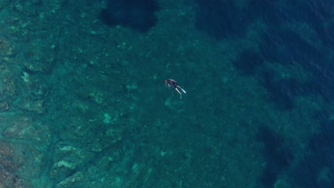 Man-snorkeling-in-circles,-crystal-clear-water,-rocky-beach-aerial-view