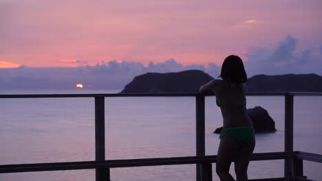 Silhouette-of-girl-in-Bikini-looking-out-on-setting-sun-over-ocean-with-islands-in-background-SLOW-MOTION