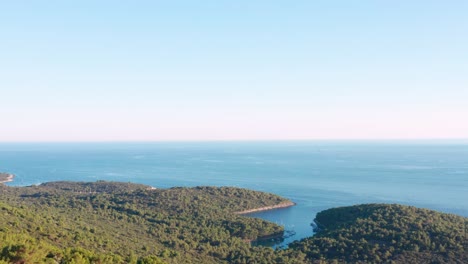 Aerial-dolly-shot-over-a-forested-ridge-revealing-the-coast-of-Losinj-island,-Croatia-below