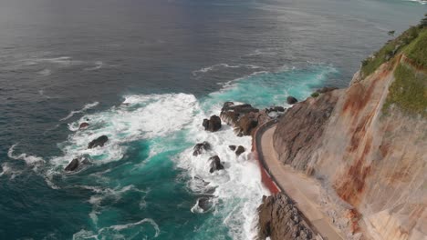 Aerial-drone-tilt-up-next-to-ocean-road-with-cliffs,-crashing-waves-next-to-beautiful-summer-island