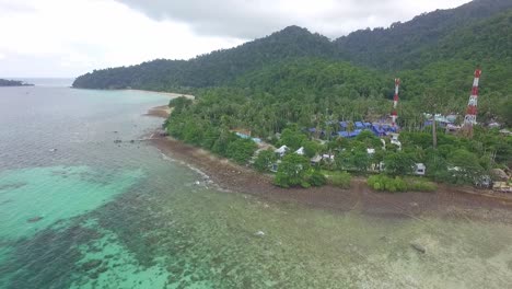 drone-shot-of-tropical-Island-with-beach,-rocky-coastline-,-small-bungalow-resorts-and-telecommunication-towers