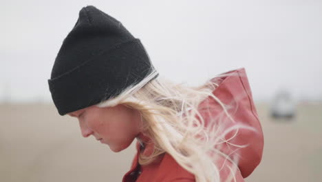Young-Girl-Wearing-Black-Beanie-And-Red-Hooded-Jacket-Looking-Down-While-The-Wind-Blows-Her-Blonde-Hair-In-Muiden,-Netherlands