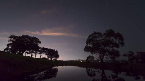Ein-Zeitraffer-Der-Sterne-über-Einer-Ländlichen-Australischen-Landschaft-Mit-Gummibäumen-Im-Vordergrund-Und-Der-Spiegelung-Des-Nachthimmels-Im-Wasser-Eines-Staudamms