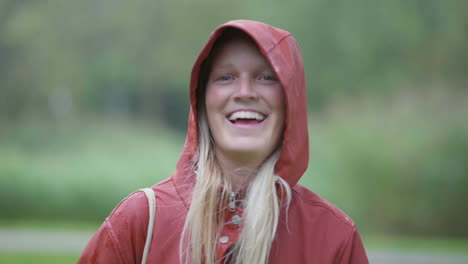 Junges,-Fröhliches-Blondes-Mädchen,-Das-In-Rotem-Kapuzenpullover-Lächelt-Und-Die-Natur-Im-Hintergrund-Verschwommen-Sieht,-In-Den-Niederlanden