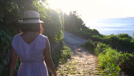Girl-in-summery-outwith-wearing-hat-walking-down-path-towards-ocean-with-sun-flaring-SLOW-MOTION