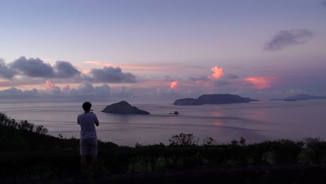 Male-traveler-taking-pictures-with-his-smartphone-on-lookout-looking-out-on-open-ocean-and-islands-at-sunrise-with-beautiful-cloud-glow