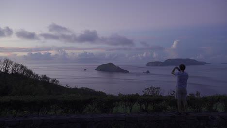 Ein-Mann,-Der-Sein-Telefon-Zum-Fotografieren-Nutzt,-Um-Fotos-Von-Einem-Aussichtspunkt-Mit-Blick-Auf-Das-Wunderschöne-Meer-Und-Die-Inseln-Zu-Machen
