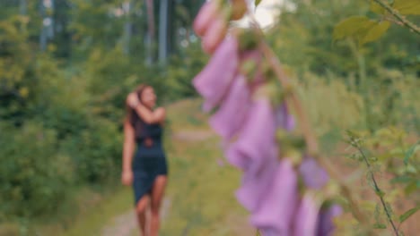 Zeitlupe-Einer-Frau-In-Schwarzem-Kleid,-Die-An-Einem-Sommertag-Auf-Einem-Natürlichen-Weg-Mit-Violetten-Fingerhutblüten-Und-Grünem-Wald-Geht,-Wechselnde-Tiefenschärfe,-Digitalis