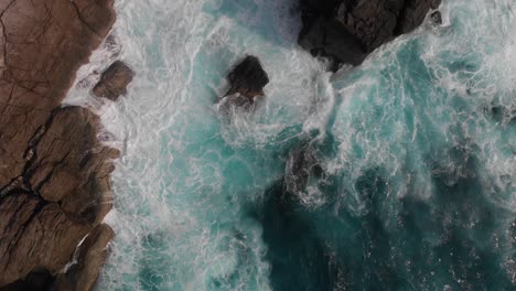 Top-down-aerial-rising-shot-over-strong-ocean-waves-breaking-against-cliffs