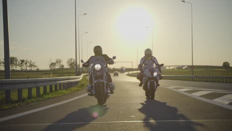 Zwei-Motorradfahrer,-Die-Verschiedene-Typen-Von-Motorrädern-Fahren,-Schlagen-Sich-Die-Hände,-Während-Sie-Bei-Sonnenuntergang-Nebeneinander-Auf-Einer-Leeren-Autobahn-Fahren