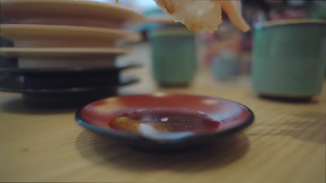 Persona-Sumergiendo-Un-Trozo-De-Sushi-Toro-De-Salmón-En-La-Salsa-De-Soja-Usando-Palillos-De-Madera-Con-Una-Pila-De-Platillo-Vacío-En-El-Fondo-Del-Restaurante-De-Sushi-En-Numazu,-Shizuoka,-Japón