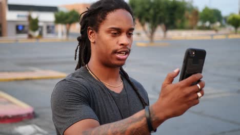 African-American-man-with-dreadlocks-and-tattoos-pulls-phone-out-to-video-call-in-parking-lot