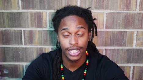African-American-man-with-dreadlocks-sitting-on-ground-looking-into-camera-saying-“no”,-“yes”,-and-“maybe”,-shaking-his-head-and-smiling
