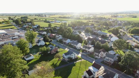 Aerial-pullback-reveals-small-rural-town-community-development,-nearby-farmland-in-Pennsylvania,-USA,-United-States-neighborhood-during-summer-magic-hour