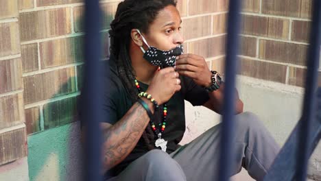 African-American-man-with-dreadlocks-sitting-on-ground-then-pulls-COVID-19-mask-out-and-putting-in-on,-then-waving-with-his-phone,-and-getting-up-to-leave
