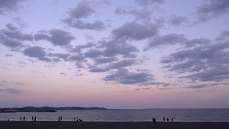 Weit-Offener-Blick-Auf-Den-Wunderschönen-Strand-Bei-Sonnenuntergang-Mit-Hohen-Wolken-Und-Mond-Und-Silhouetten-Von-Menschen,-Die-Im-Sand-Spazieren-Gehen