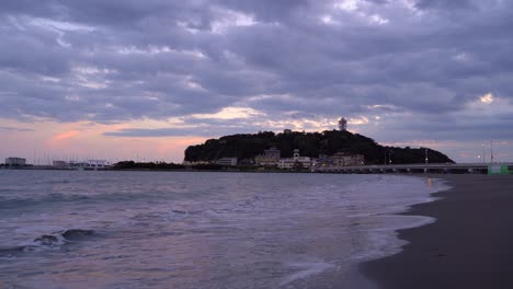 Tiefwinkel-Weitblick-Auf-Die-Insel-Enoshima-In-Japan-Bei-Sonnenuntergang-Mit-Wellen,-Die-Am-Strand-Brechen
