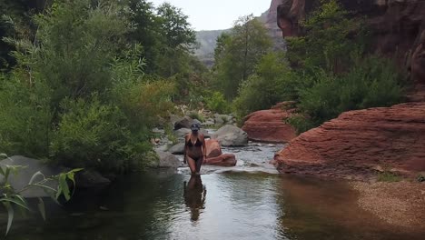 Model-in-bikini,-slow-motion-walking-out-of-running-water-river-surrounded-by-rocks-and-trees