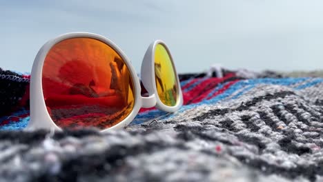 Close-up-of-red-tinted-reflective-white-sunglasses-with-the-reflection-of-a-woman-sunbathing-on-a-beach-blanket
