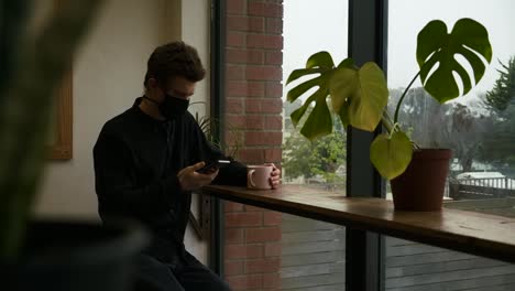 Young-adult-man-sitting-in-restaurant-using-smartphone-wearing-covid-prevention-mask