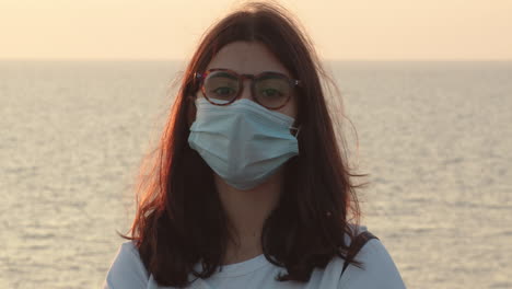 Front-view-of-young-European-girl-wearing-mask-looks-into-camera-at-sea-background,-hopeless-about-future-of-pandemic-covid-19,-static,-close-up