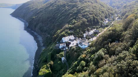 Bucks-Mills-Aerial-drone-flys-along-wooded-Devon-coast-line-small-village-on-left-of-frame-Sunny-day-beach-in-shadow