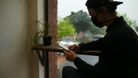 Young-adult-man-sitting-in-coffee-shop-window-on-smartphone-wearing-covid-mask
