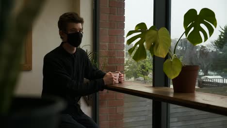 Young-adult-man-sits-looking-out-of-restaurant-wearing-covid-prevention-mask