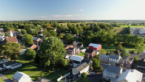 High-and-wide-aerial-overhead-shot-of-small-American-town,-village,-establishing-shot-shows-homes-and-housing-along-street,-green-trees-and-farmland-in-distance-during-August,-North-America,-USA