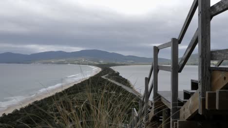 Wind-blows-bushes-overlooking-long-island-isthmus-surrounded-by-ocean-beach-waves-and-green-mountain-and-forest-on-overcast-cloudy-day-as-car-drives-down-road,-Bruny-Island,-Tasmania,-Australia