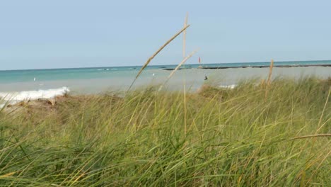 Nahaufnahme-Von-Strandgräsern,-Die-Sanft-Im-Wind-Wehen---Mit-Wasser,-Surfer-Und-Leuchtturm-Im-Hintergrund