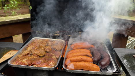 Grilling-delicious-meat-sausages-and-steaks-on-smoky-grill-with-flames-on-foil-trays
