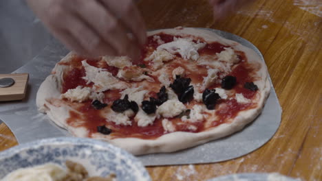 Chef-tops-Neapolitan-pizza-with-black-olives,-on-wooden-table