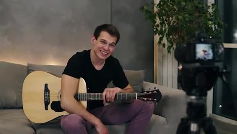Handsome-caucasian-man-in-black-T-shirt-making-video-blog-about-musical-instruments-or-recording-the-song-for-his-followers-while-sitting-on-a-couch-at-home.-Recording-camera-on-a-foreground