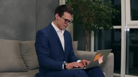 Caucasian-businessman-wearing-formal-blue-suit-and-stylish-glasses-sitting-on-the-sofa-typing-on-his-laptop-on-knees-at-the-hotel-room.-Loft-interior-room
