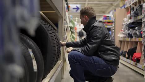 Young-man-in-supermarket-choosing-automobile-tyres-standing-on-the-shelves